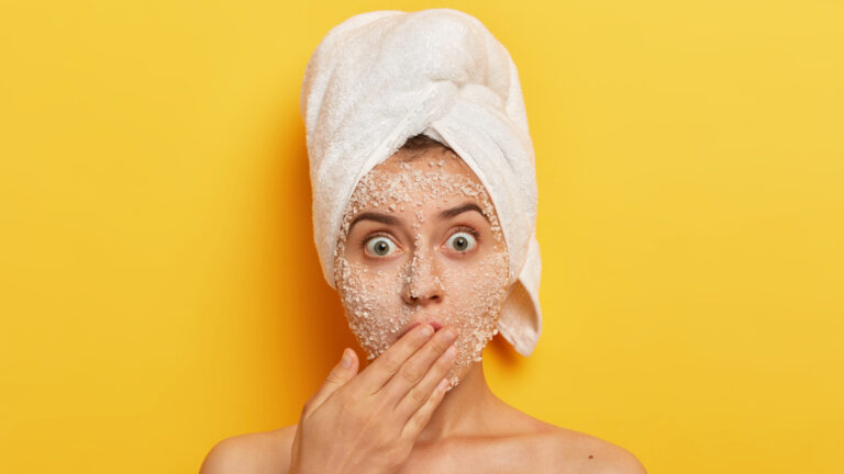 Headshot of stupefied young woman with natural scrub mask on face, gasps from fear, covers mouth with palm, reduces pimples stands shirtless exfoliates blackheads as she has applied face scrub