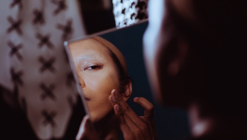 A lady applying watery foundation with her fingers and looking at herself in a square mirror