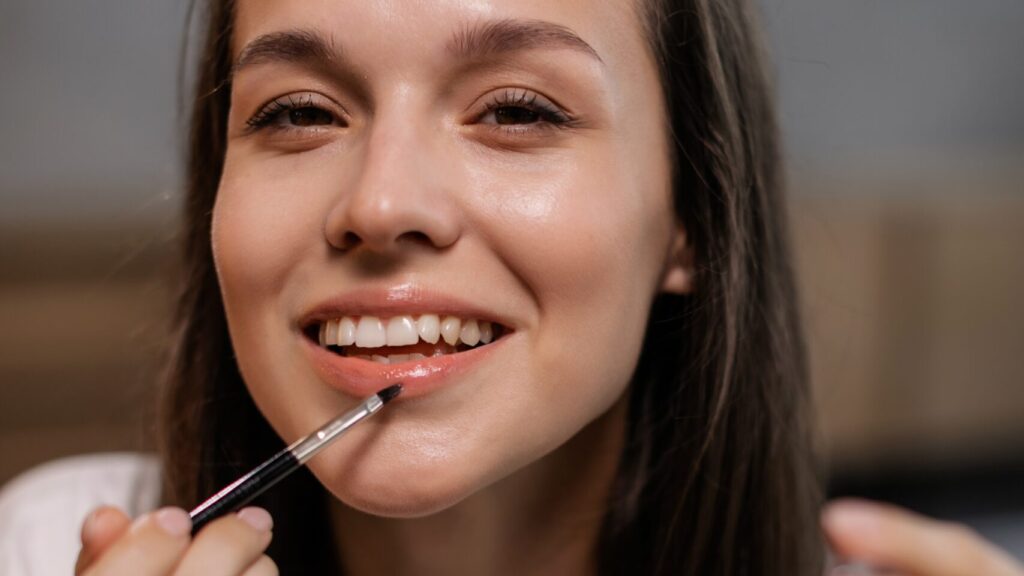 a woman applying lip gloss as she smiles into the camera