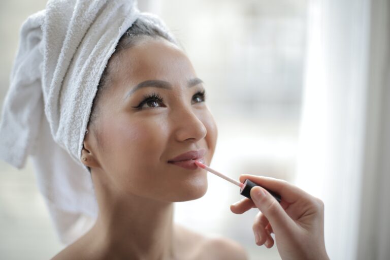 a woman with towel standing while a hands applies lip gloss to her lips