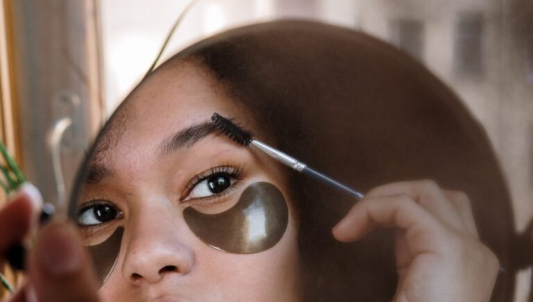 reflection of an afro woman with under eye mask, brushing through her eyebrows to see her eyebrow filler.