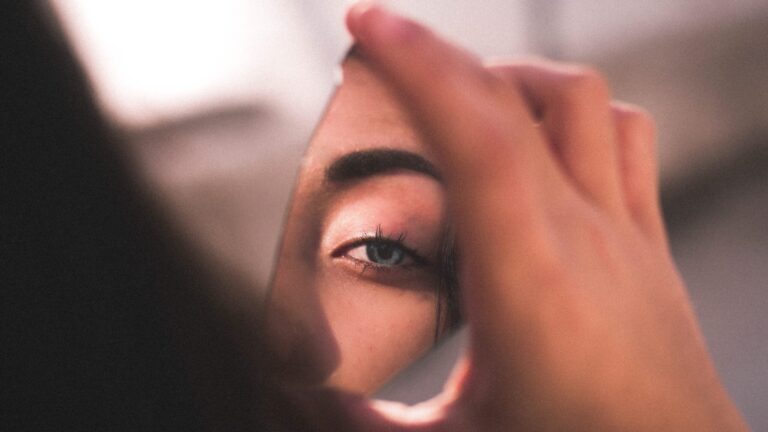 a woman staring into a broken piece of mirror she is holding in her hand as she has face serum on