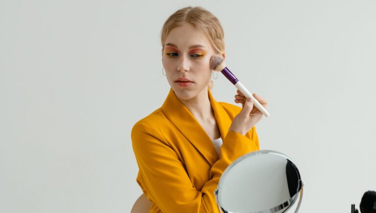 woman in yellow suit applying makeup with brush as she looks into a mirror.