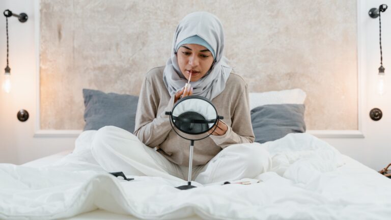 a woman in hijab sitting in bed looking into a mirror as she applies lip tint