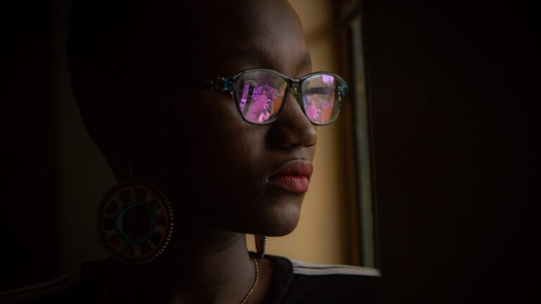 an afro woman with glasses looking out of a window as she has lip gloss on