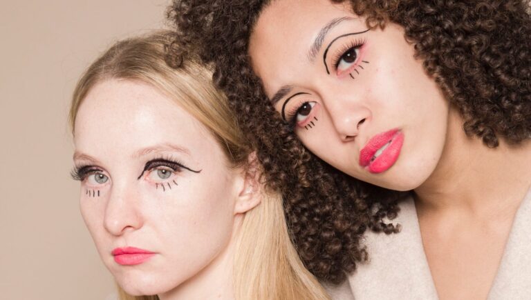 two women with black eyeliner posing for a picture, one is wearing off white tee