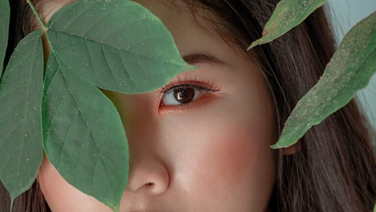 an Asian woman posing behind green leaves as she's showing off her permanent eyeliner