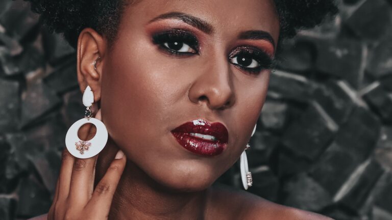 an afro woman with red lip gloss and brown eyeshadow standng in front of leaves