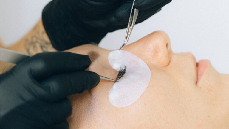 woman laying on a bed as two hands with black gloves are doing lash lift procedure on her