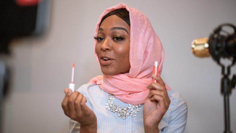 an afro woman with pink scarf holding a clear lip gloss for dark skin