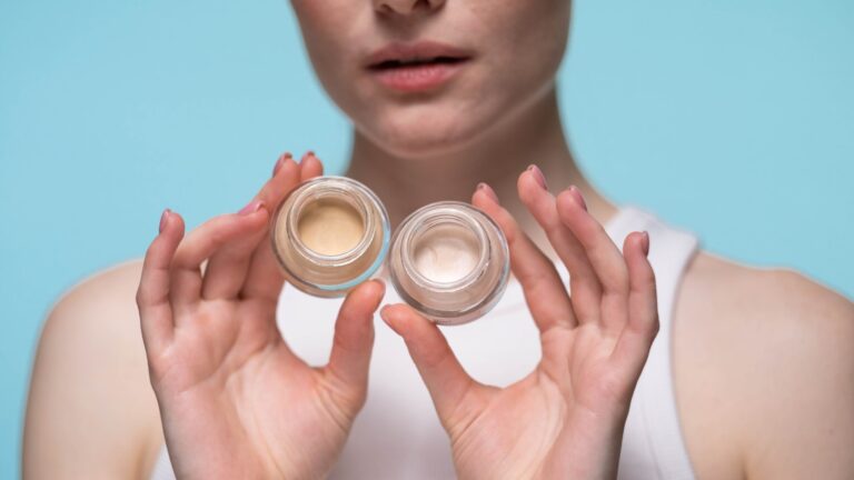 woman holding two concealer holders in her hands as she stands in front of a blue background wearing a white tank top