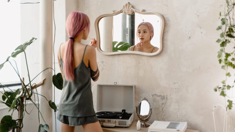 a woman with pink hair and charcoal clothes standing in a room with plants and looking into a mirror as she applies lip gloss