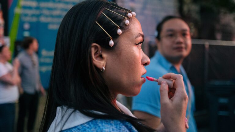 a woman looking side ways as she applies lip tint to her lips, a man stands behind her staring at her