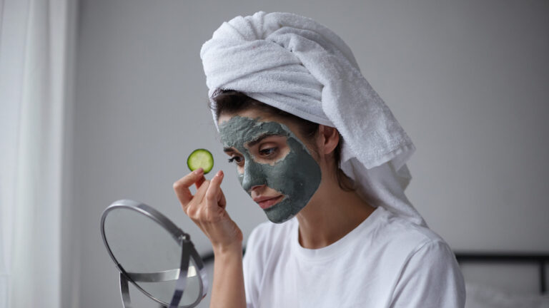 Beauty portrait of attractive young dark haired calm female in chemical body scrub with fresh cucumber in her hand looking into mirror, keeping lips folded.