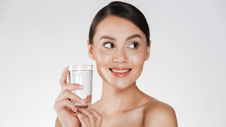 Beauty portrait of young happy woman with hair in bun holding micellar water from transparent glass isolated over white background