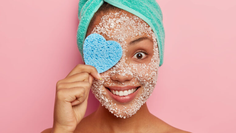 Headshot of cheerful young woman covers one eye with heart shaped cosmetic sponge, removes makeup, has sea salt scrub around face, tender smile, happy expression, cares about complexion and skin