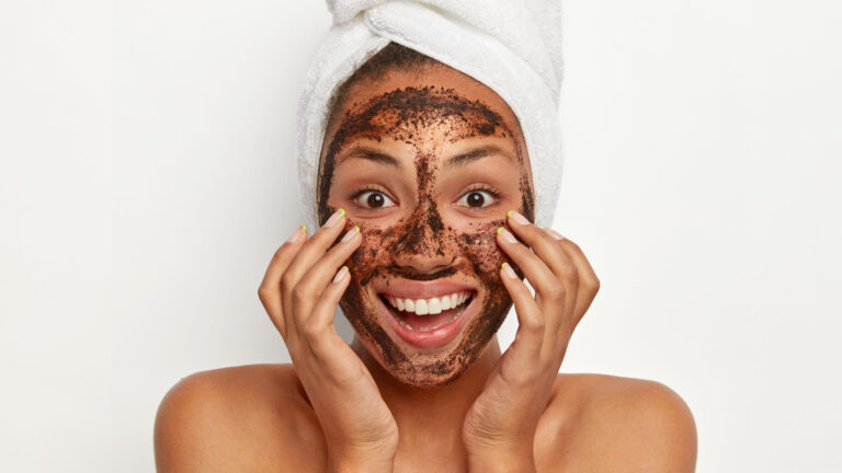 Headshot of beautiful dark skinned woman wears coffee scrub face scrub, keeps hands on cheeks, smiles broadly, wears white towel on head, models indoor against white background.