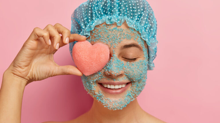 Headshot of cheerful young woman with crystal sea salt face scrub, holds pink heart shaped sponge on eye, smiles positively, wears showercap, models against pink background.