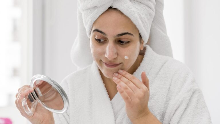 medium shot of a woman wearing a robe and a towel over her head and applying a chemical body scrub on her face while looking at a mirror