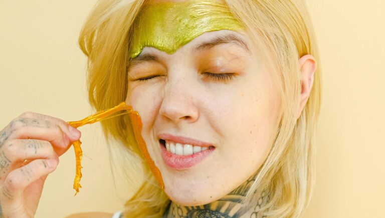 a white light skinned woman with tattoos on her neck and hand peeling turmeric face mask while she has turmeric face scrub on her forehead.