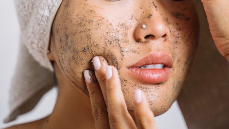 A woman wearing a white towel on her head wearing a nose pin, touching her face that has face scrub on it.