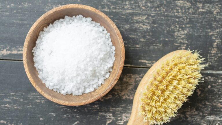 a bowl of salt scrub besides a brush on a marble surface