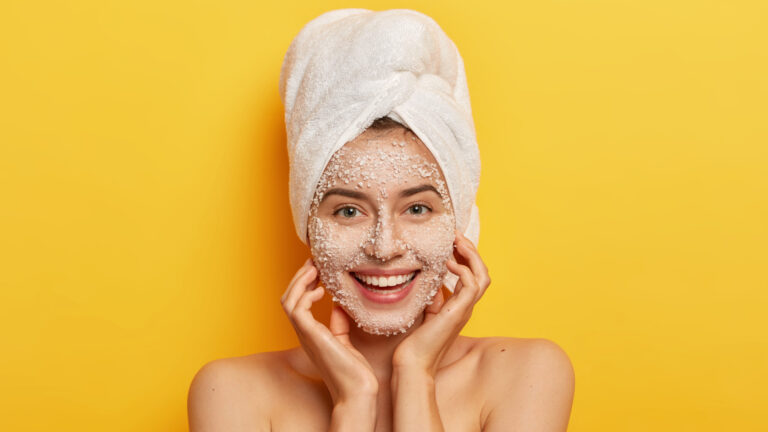 Pleasant looking happy woman unclogges pores, makes beauty step to improve skin, wears nourishing facial scrub, supples complexion, keeps hands on cheeks, stands naked against yellow studio wall