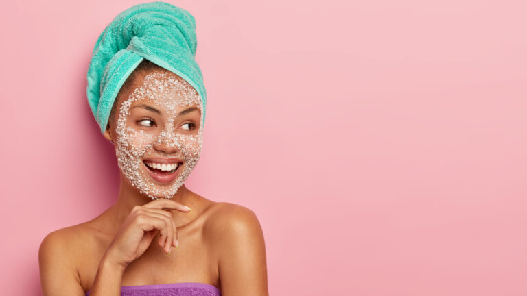 woman wearing a turquoise towel looking at her right side and touching her face with her knuckles as she has face scrub on.