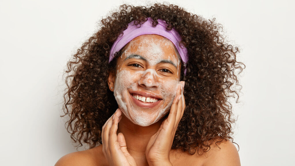 Skin care and ethnicity concept. Beautiful satisfied curly woman washes face, has foam on skin, looks happily at camera, stands topless against white background, enjoys freshness and cleanliness as she applies body scrub on her face