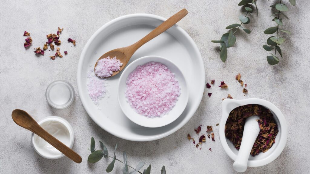 pink salt scrub in a bowl placed in a white plate with a wooden spoon. there are some green plants and two more bowls of spices besides the pink salt