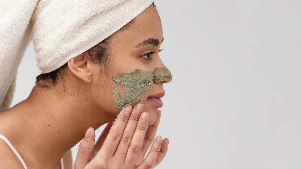 a woman applying face scrub of gray color with her hands in front of a grey background