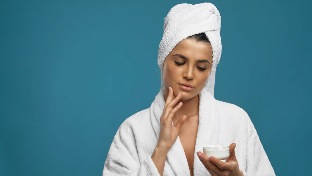 Front view of young woman in bathrobe and towel on head using cream for hydrating and rejuvenating of skin. Portrait of pretty female after shower, isolated on blue. thinking of applying face scrub on her face