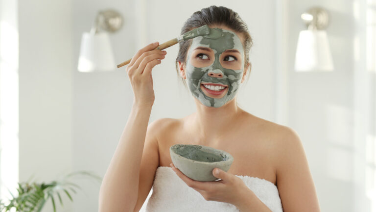 A woman wearing white towel, holding a mud bowl with face scrub in it, as she's applying it on her face with a brush. she is smiling