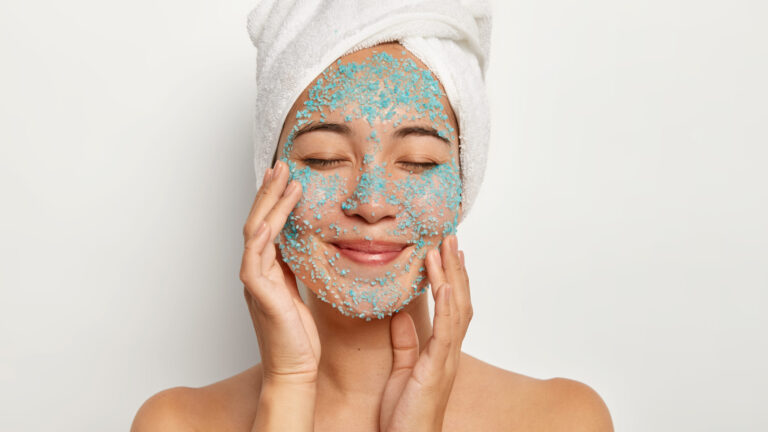 a woman smiling as she's touching her face with grey face scrub on it